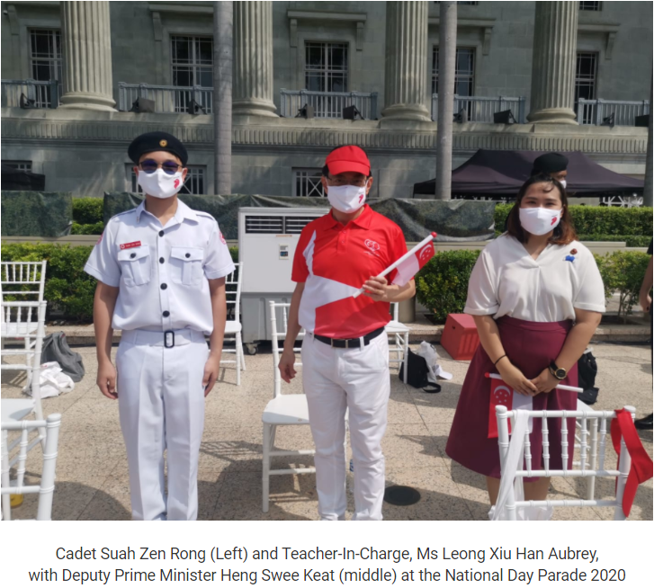 Three masked people stand outdoors at the 2020 National Day Parade, wearing white uniforms and red attire.